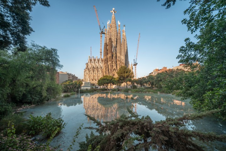 Sagrada Familia reflected in a pond, surrounded by trees and cranes.