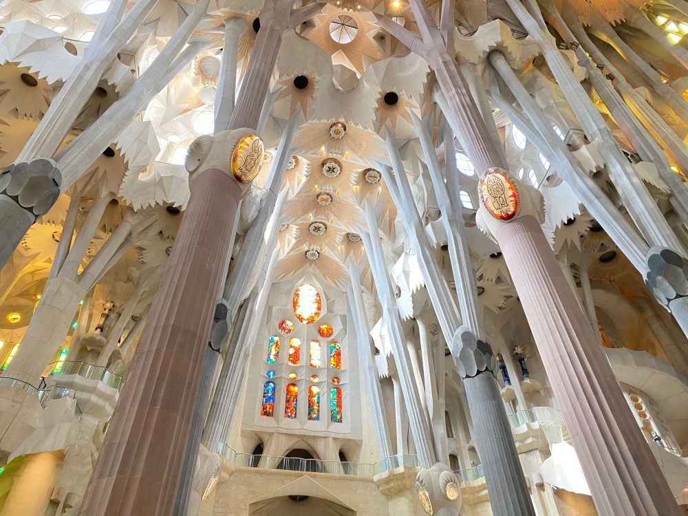 Looking up at the tree-like columns and intricate vaulted ceiling of the Sagrada Familia, with colorful stained glass windows in the background.