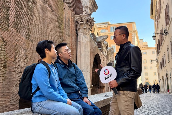Two people sitting on a ledge listening to a man holding a microphone in a historic street.