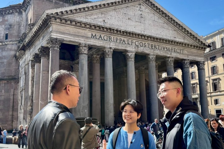 Three people talking in front of the Pantheon, Rome, with crowds in the background.