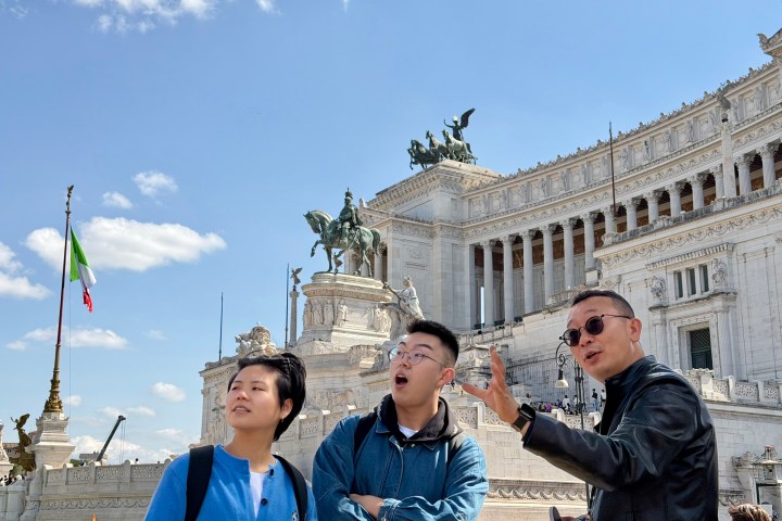 Three people in front of a historic building with statues and an Italian flag.