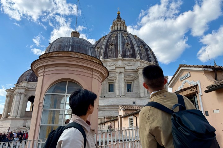 Two people with backpacks admiring a large domed building with blue sky and clouds.
