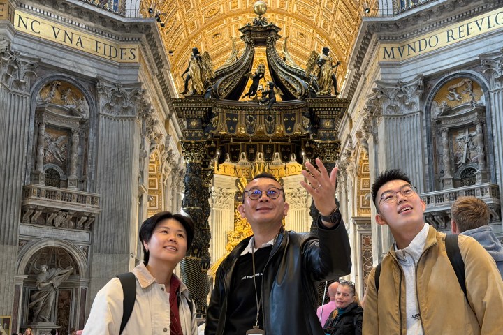 Three people look up inside an ornate cathedral, with a decorative black canopy overhead.