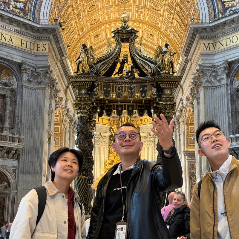 Three people look up inside an ornate cathedral, with a decorative black canopy overhead.