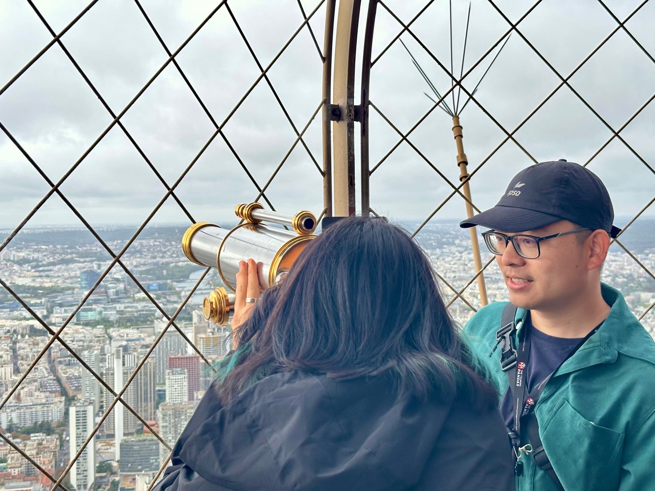 Two people on a tower balcony, one looking through binoculars, with a city view in the background.