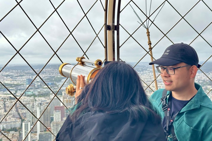 Two people on a tower balcony, one looking through binoculars, with a city view in the background.