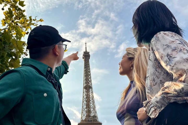 Three people admire the Eiffel Tower, with one pointing towards it under a partly cloudy sky.