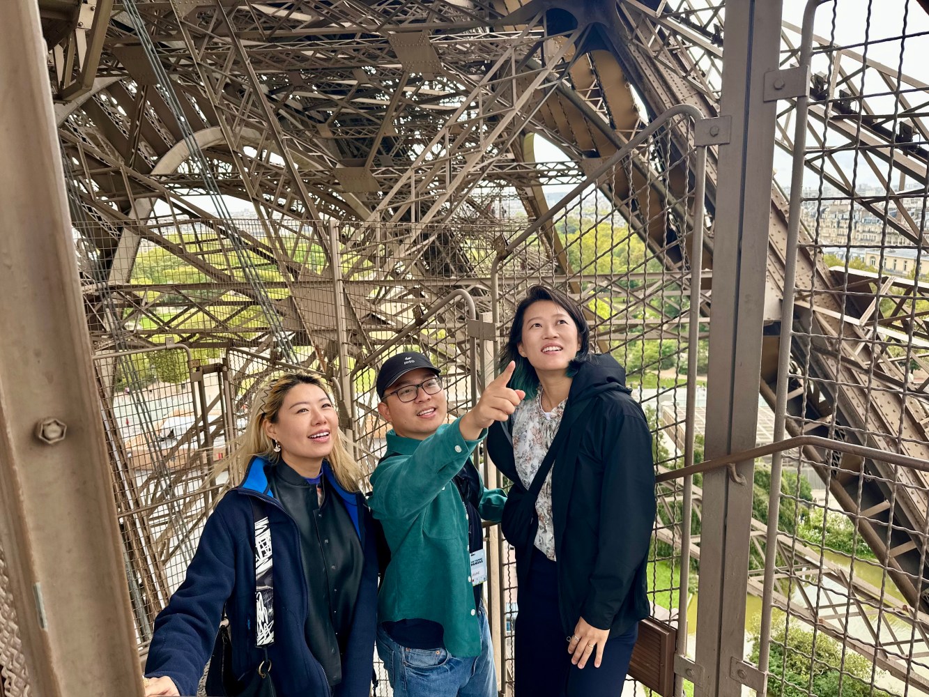 Three people on Eiffel Tower platform, one pointing upward, surrounded by metal structure.