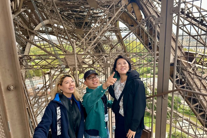 Three people on Eiffel Tower platform, one pointing upward, surrounded by metal structure.