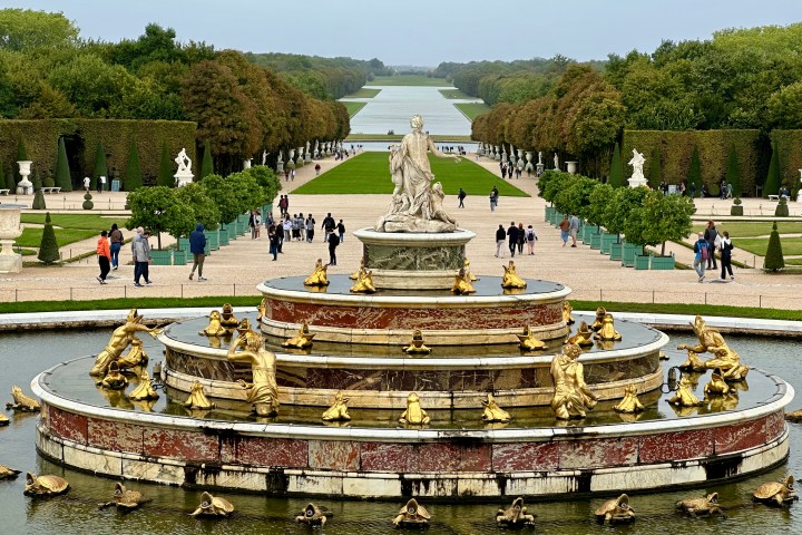 Ornate fountain with golden statues in a formal garden, people walking along paths lined with trees.