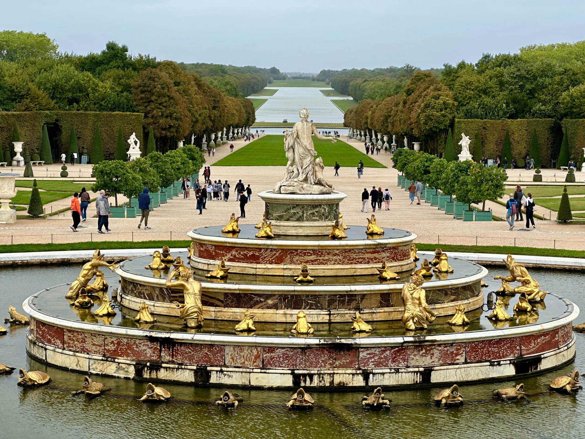 the-touring-pandas-versailles-tour-8 Ornate fountain with golden statues in a formal garden, people walking along paths lined with trees.