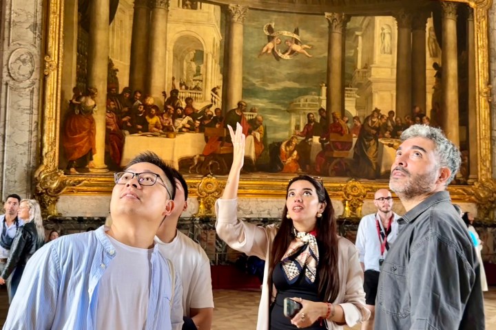 Three people in a museum admire a large, detailed painting with gold frame.