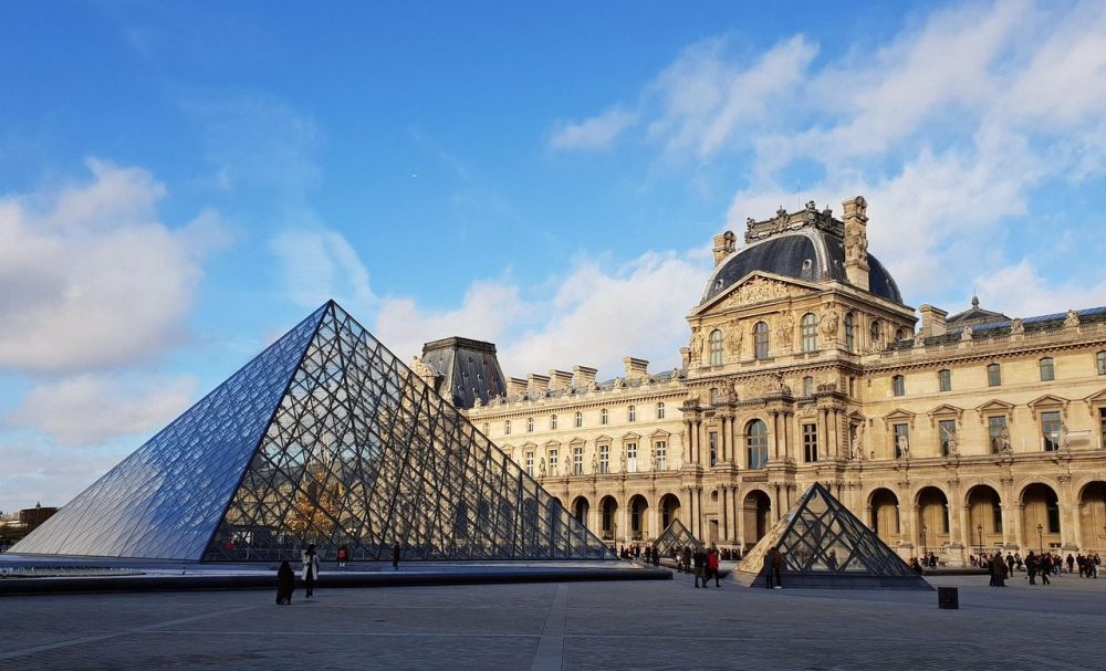 Glass pyramid in front of historic building with blue sky and clouds.