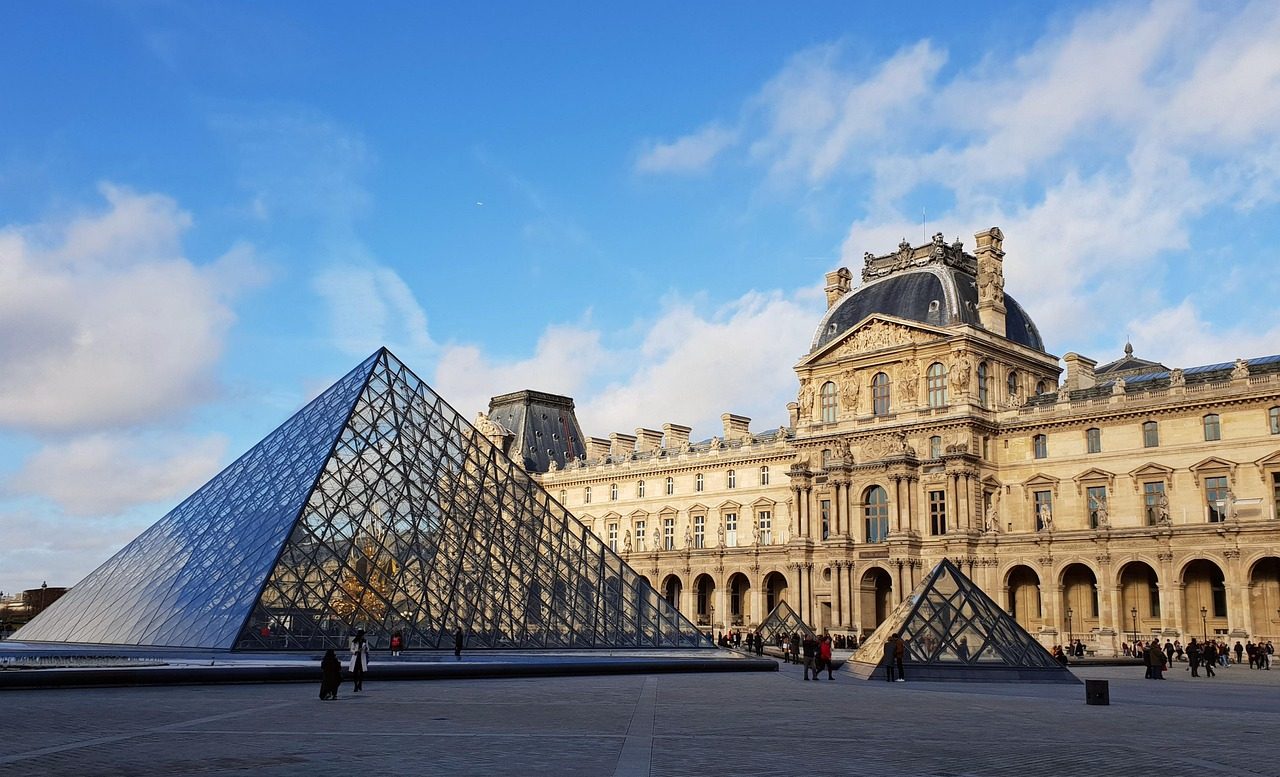 louvre-5767708_1280 Glass pyramid in front of historic building with blue sky and clouds.