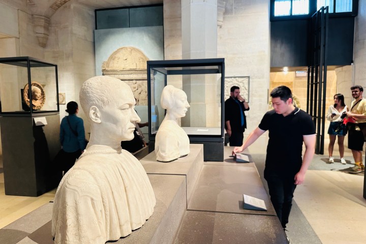 Museum visitors view ancient busts displayed in a gallery with stone walls.