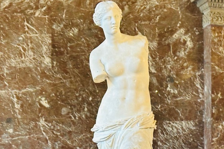 Marble statue of a female figure with missing arms against a stone wall.