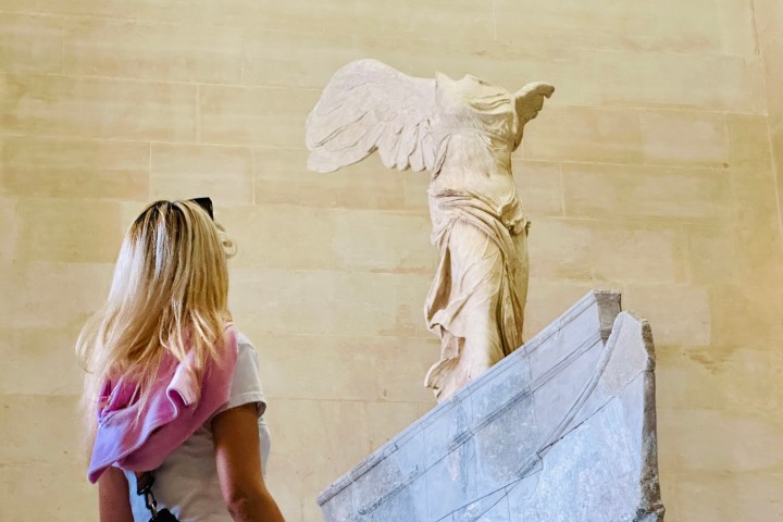 Person with long hair views the Winged Victory of Samothrace statue in a museum.
