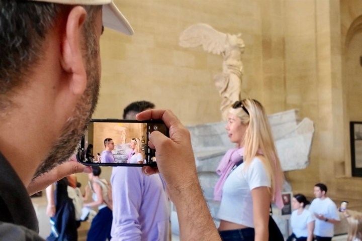 Person photographs Winged Victory statue in museum with phone, capturing tourists in the scene.