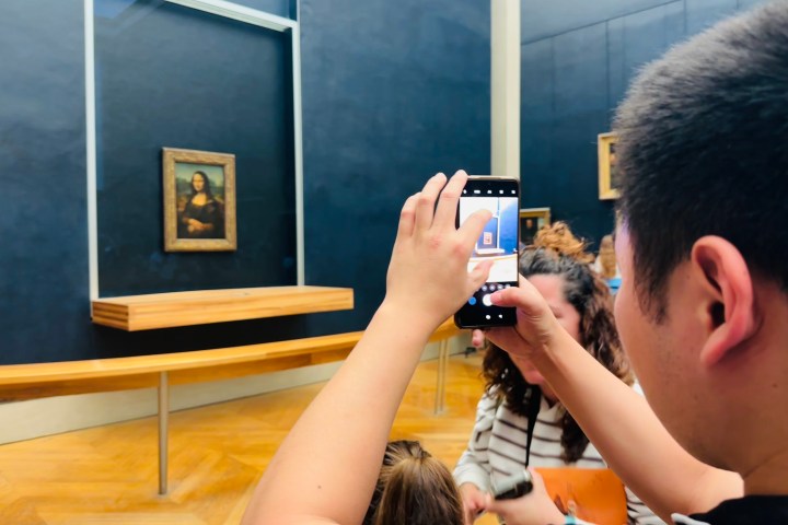 People photographing the Mona Lisa inside the Louvre Museum using smartphones.