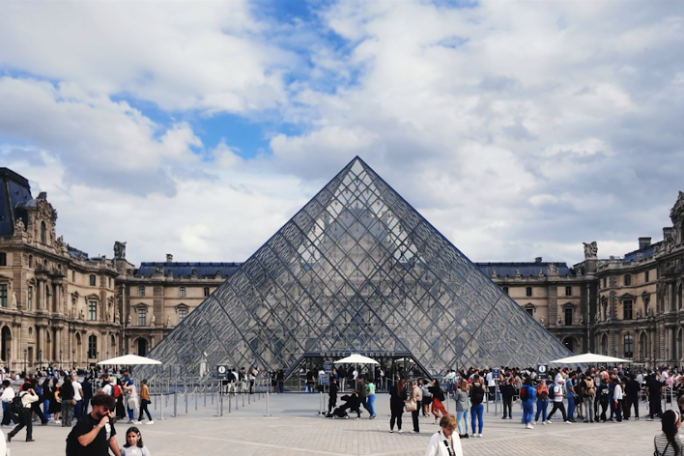 Visitors at the Louvre Pyramid entrance under a partly cloudy sky.