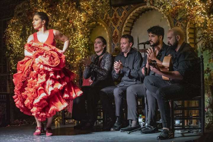Flamenco dancer in red dress performs on stage with four musicians seated, clapping.