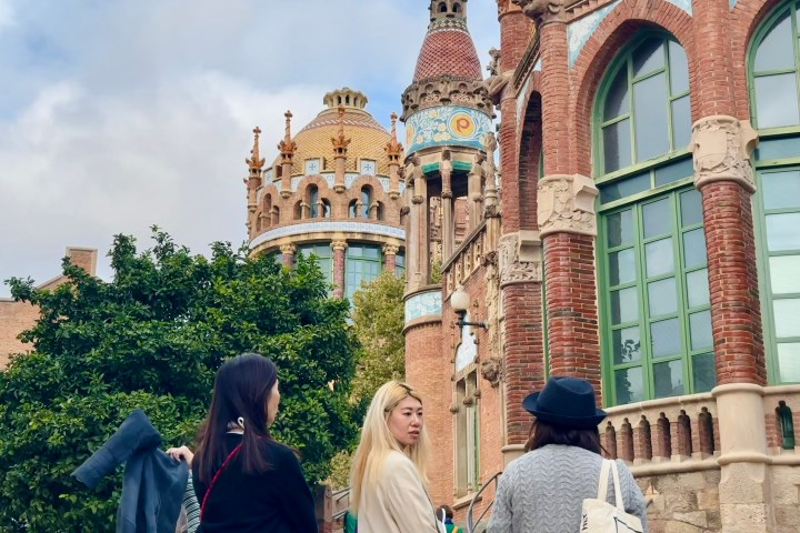 Three people walking near a historic brick building with ornate towers and archesduring the sant pau art nouveau site guided tour