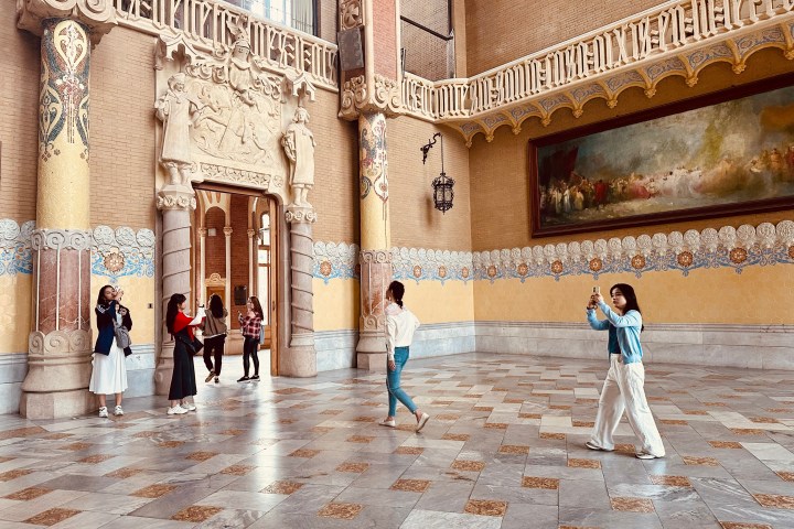 Ornate hall with people taking photos, featuring decorated columns and a large painting. Taken during the sant pau art nouveau site tour.