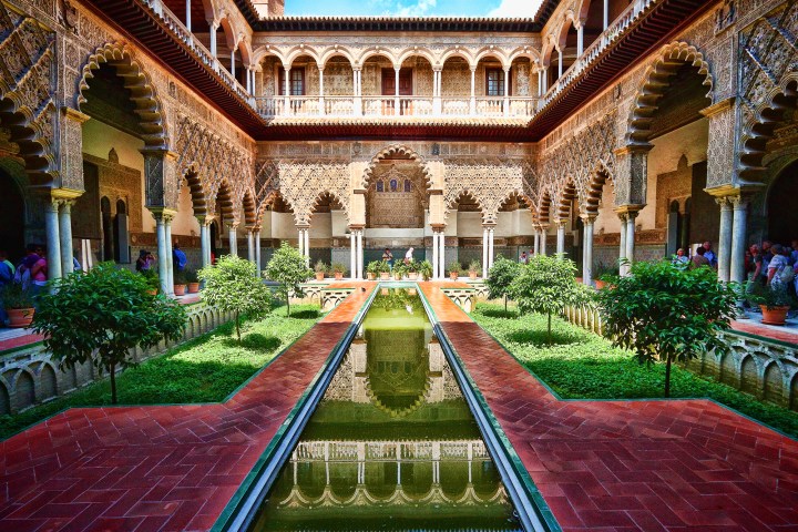 Ornate courtyard with arches, reflecting pool, symmetrical gardens, and people admiring the architecture.