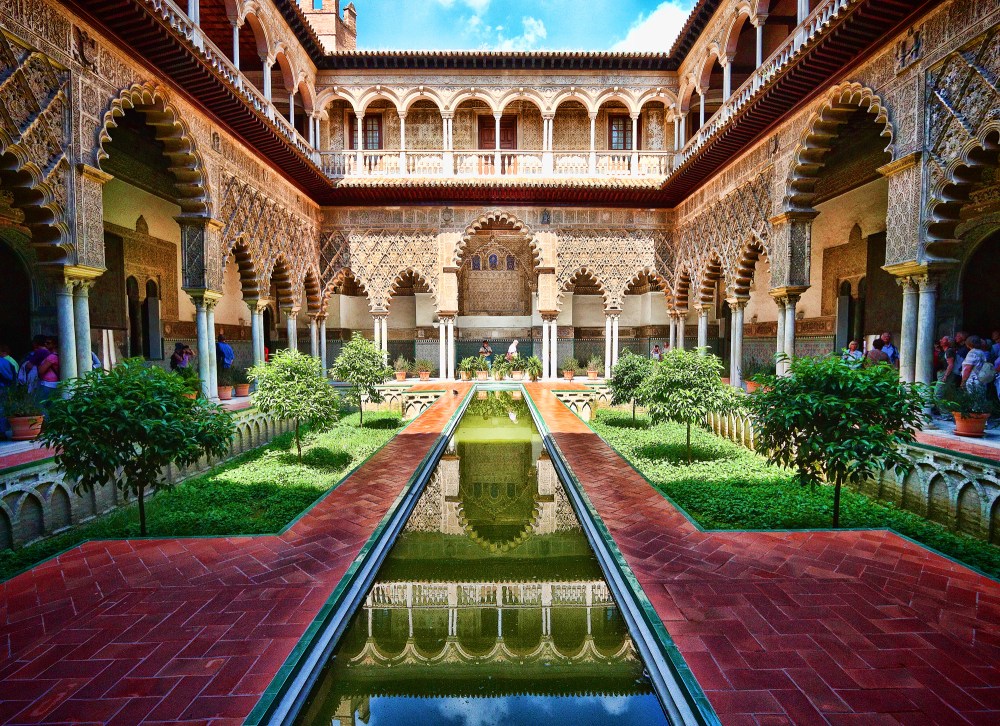 Ornate courtyard with arches, reflecting pool, symmetrical gardens, and people admiring the architecture.