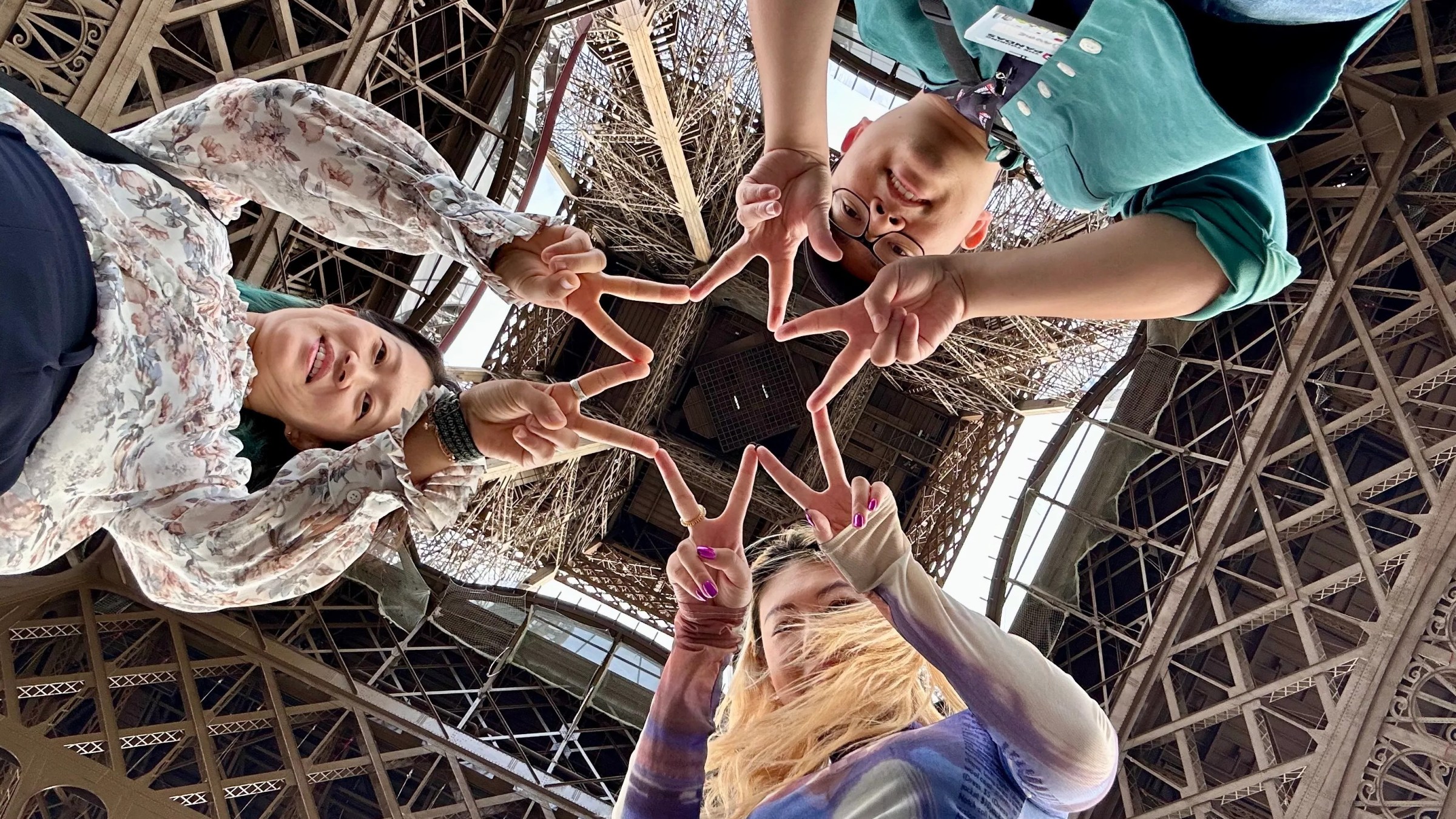 Four people making a star shape with fingers, looking up under the Eiffel Tower.