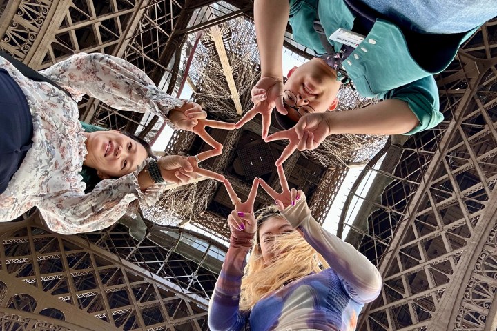 Four people making a star shape with fingers, looking up under the Eiffel Tower.