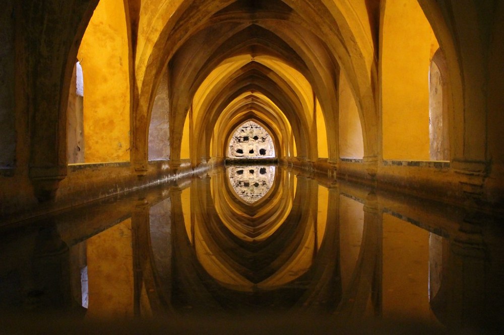 Arched hallway with warm lighting reflected on a still water surface, creating a symmetrical mirror effect.