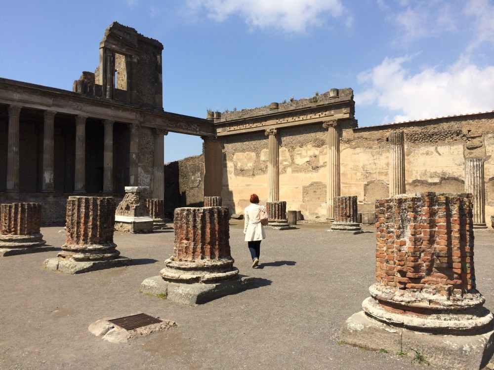 Person walking among the ancient ruins of pompeii with brick columns and a clear blue sky.