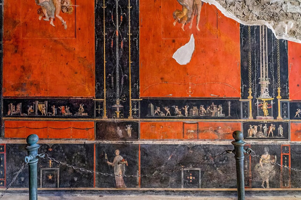 Ancient Roman fresco with red and black panels, figures, and decorative motifs on a wall.