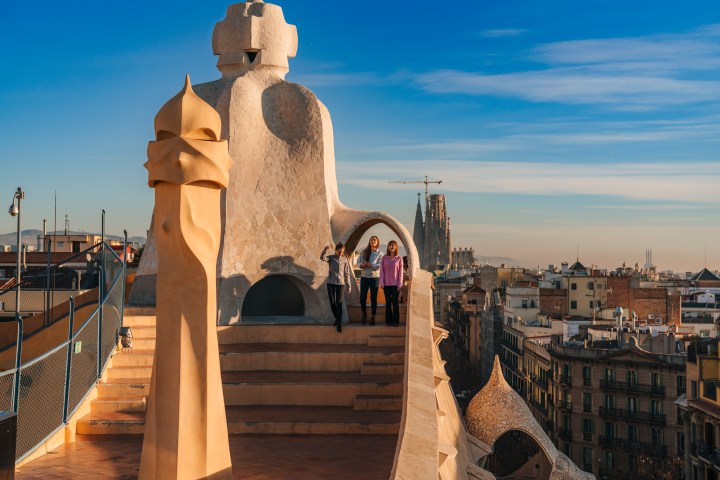 Three people on a rooftop with sculptural chimneys and a cityscape background under a blue sky.