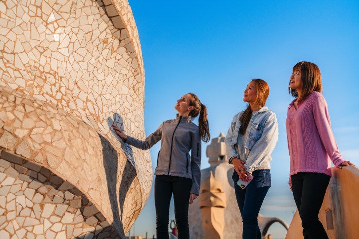 Three women observing mosaic wall on a sunny day with blue sky.