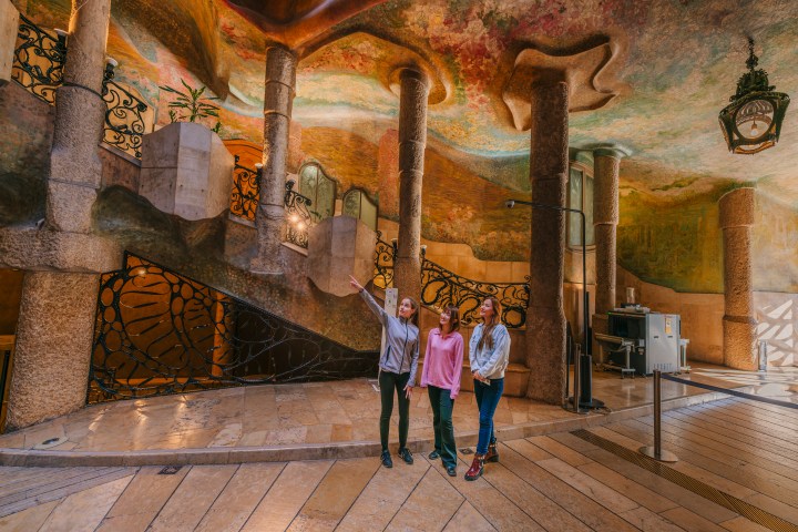 Three people standing in an ornate hall with colorful ceiling and stone columns.