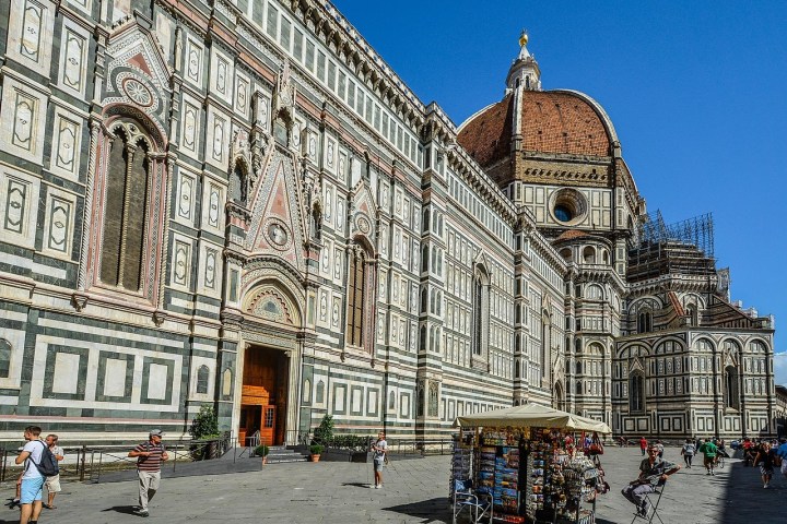Cathedral with intricate facade and large dome, tourists and souvenir stall in foreground on sunny day.