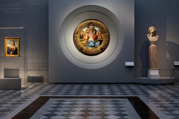 Art gallery with framed paintings and marble bust on gray walls and checkered floor.
