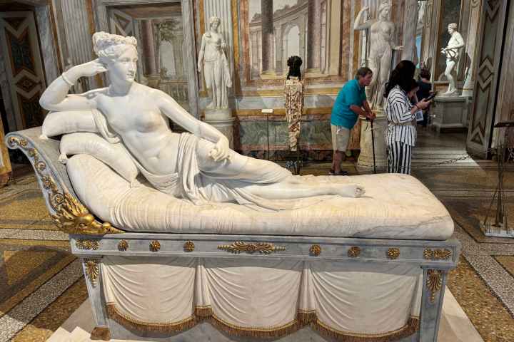 Marble sculpture of a reclining woman in a classical setting with ornate decor and people viewing.