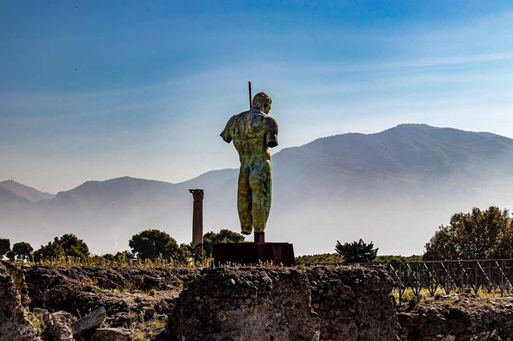 Back view of a statue overlooking Vesubio Volcano and Pompeii ruins under a clear sky.