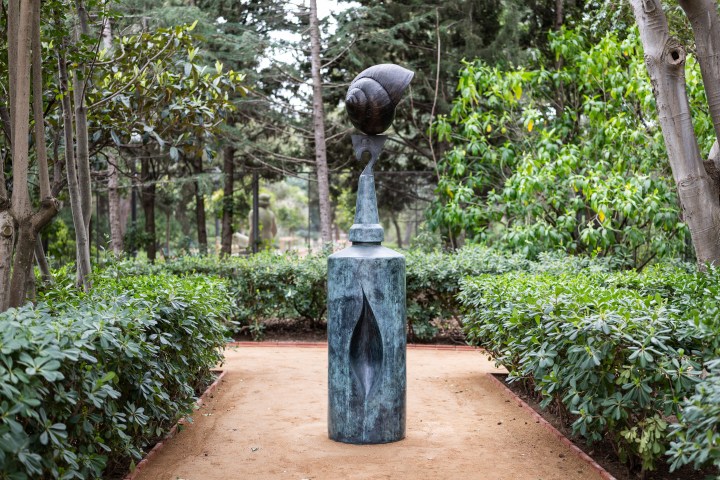 Outdoor sculpture in garden path, surrounded by trees and bushes.
