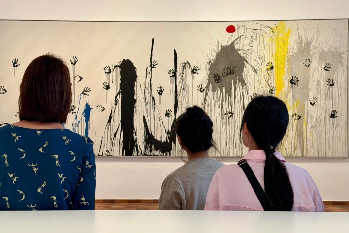 Three people view a large abstract painting with handprints and splashes of color.