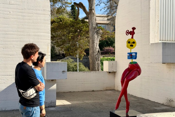 Two people observe a colorful abstract sculpture in an outdoor setting.