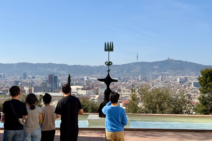 Tourists view cityscape from a terrace, featuring hills and a sculpture in the foreground.