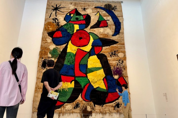 Visitors view a large, colorful abstract tapestry in a museum.