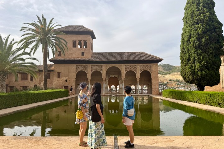 Three people stand by a reflecting pool in front of a historic building with arches and palm trees.