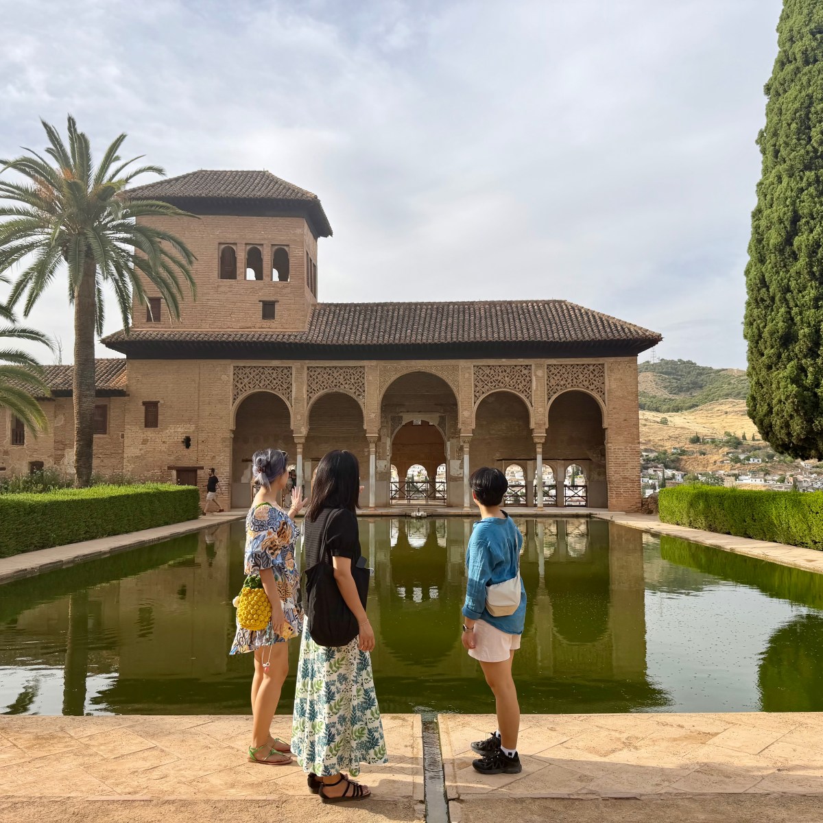 Three people stand by a reflecting pool in front of a historic building with arches and palm trees.