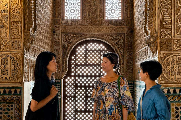 Three people talking in ornate room with intricate wall patterns and lattice window.