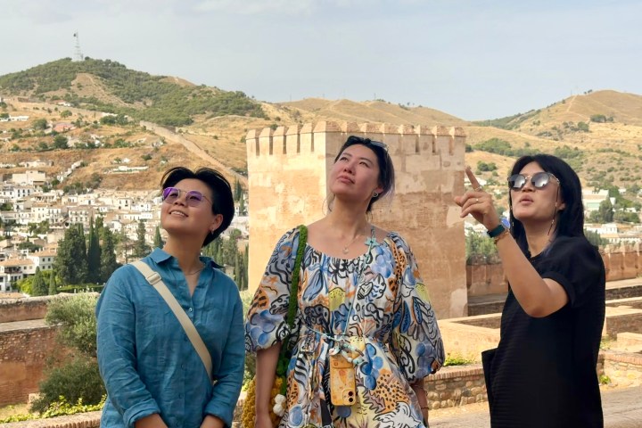 Three people outdoors, one pointing, with hills and ancient wall in the background.